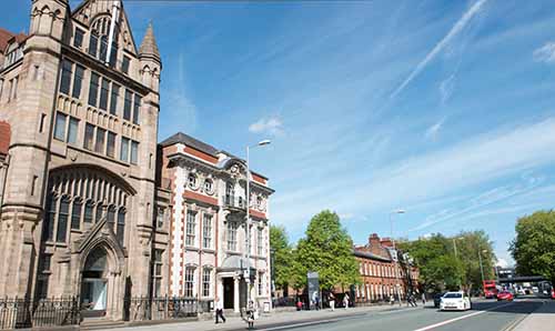 Street view of Manchester Museum, showcasing its historic stone and brick buildings, with pedestrians and traffic, including a red double-decker bus, under a bright blue sky with wispy clouds.