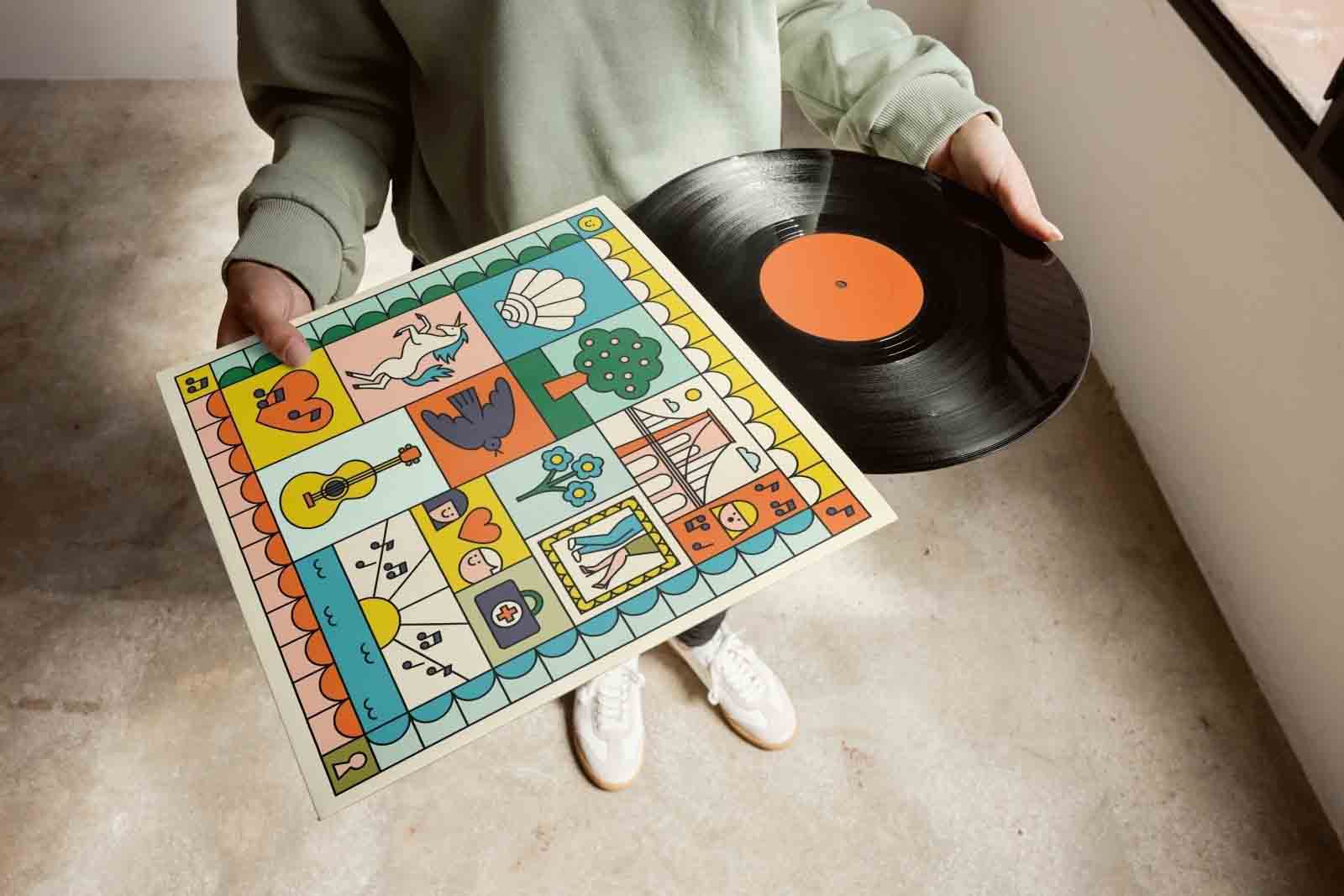 A person in a green sweatshirt holds a vinyl record and a colorful album cover with various illustrations. The setting has a concrete floor and natural light.